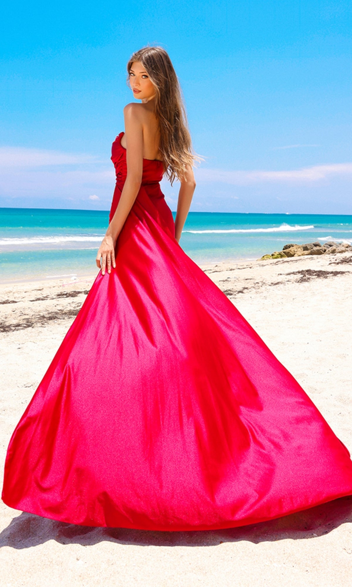 A woman stands on a sandy beach, facing the camera in the Strapless Long Formal Prom Dress with Drape 3013, with the ocean and blue sky in the background.